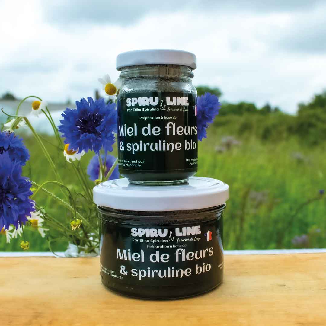 Pots de miel de fleurs et spiruline bio 125g et 250g placés devant un champ de fleurs avec des serres de spiruline en arrière-plan.