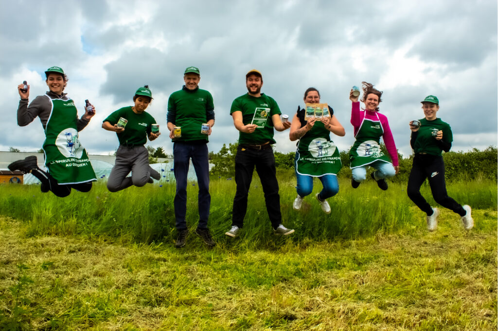 L'équipe Etika Spirulina tenant les produits à base de spiruline Etika Spirulina dans un champ fleuri, avec les serres de spiruline en arrière-plan.